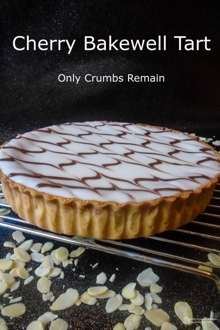 whole cherry bakewell tart on a wire rack with flaked almonds scattered on the work surface. with text overlay for pinterest.