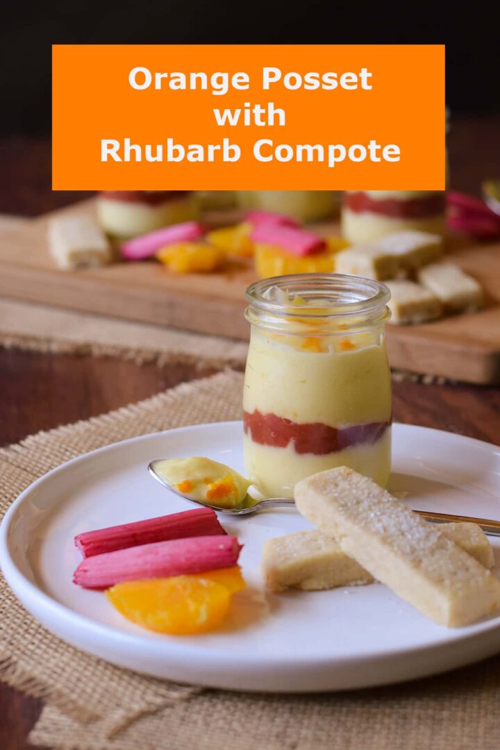 orange posset layered with rhubarb compote in glass jar with spoon ful removed. served with shortbread orange slices and rhubarb batons.