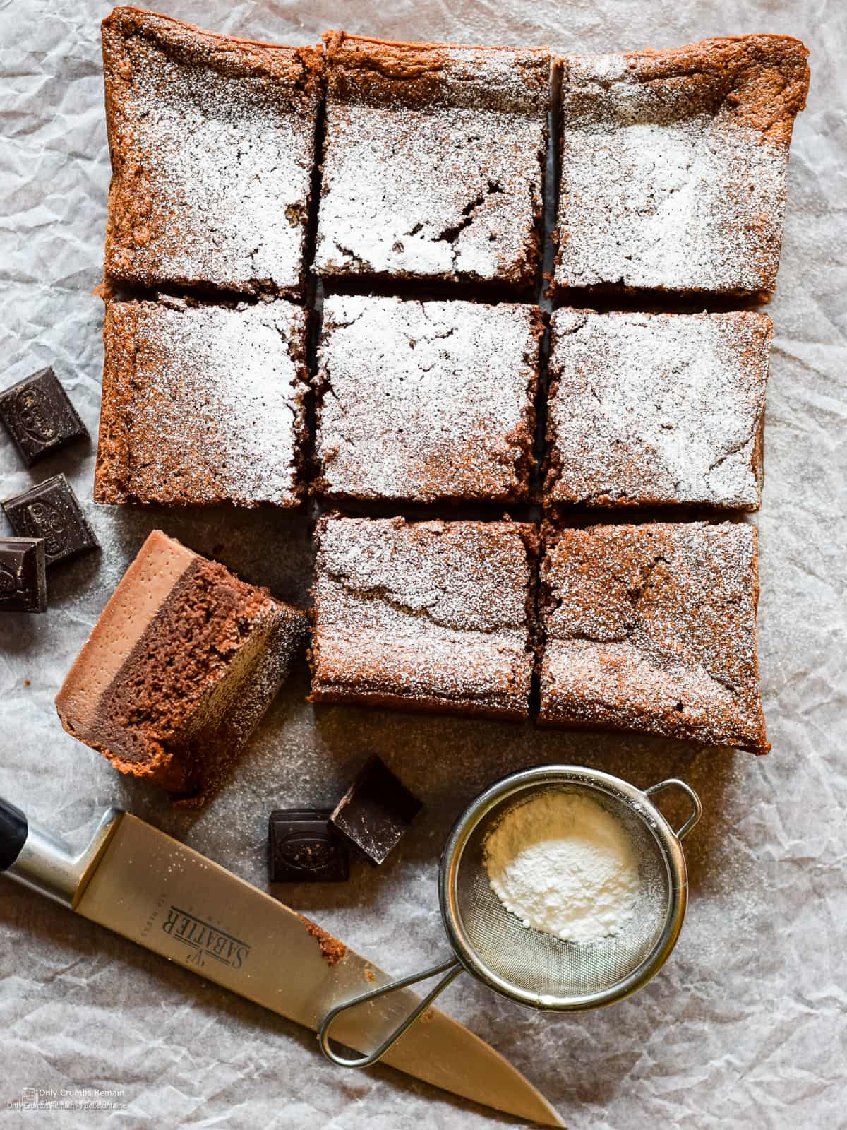 chocolate orange magic cake cut into squares with pne piece turn on its side. on baking parchment with knife, sieve of icing sugar and cubes of chocolate.