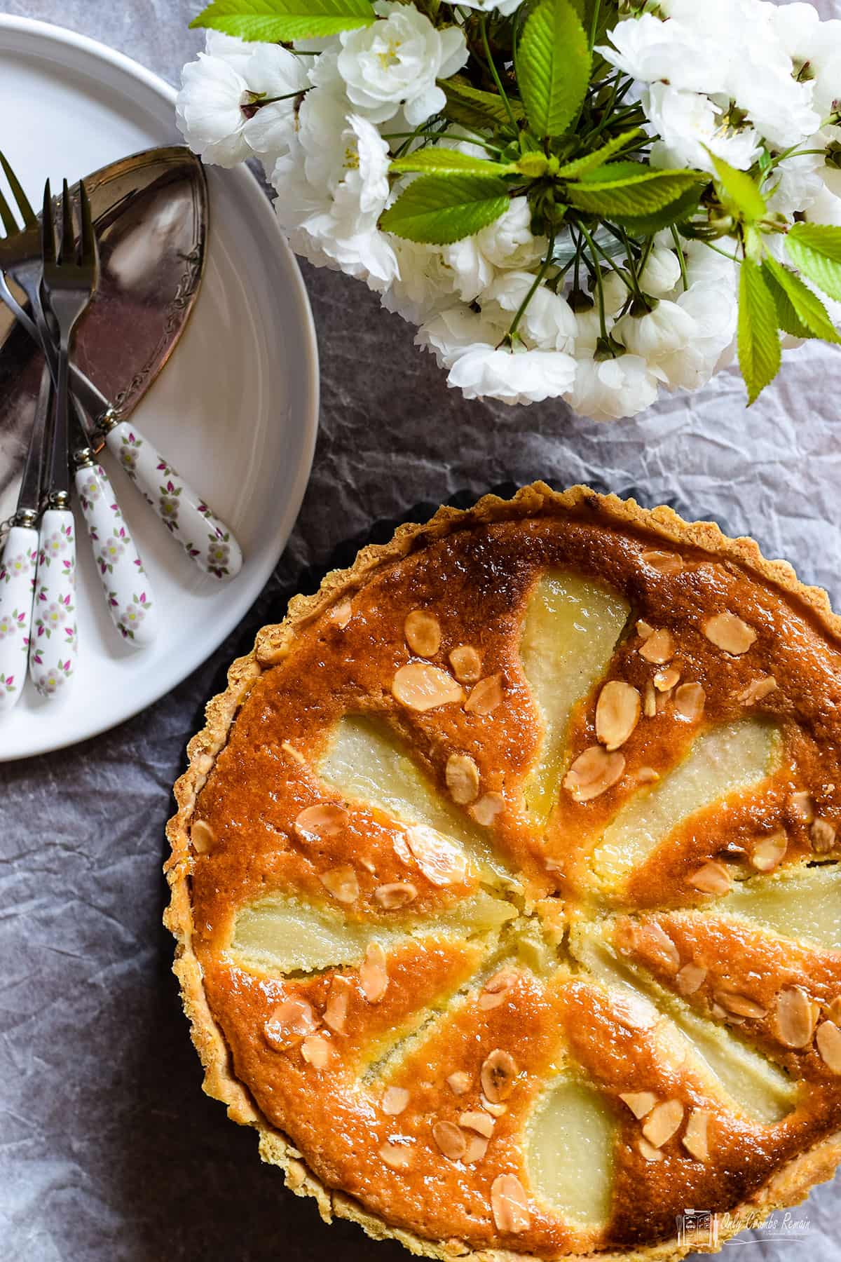 Pear and almond tart with plate of cutlery and vase flowers above.