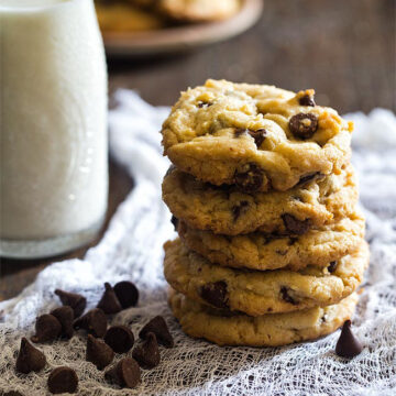 stack of chocolate chip cookies with bottle milk behind and some choc chips in front.