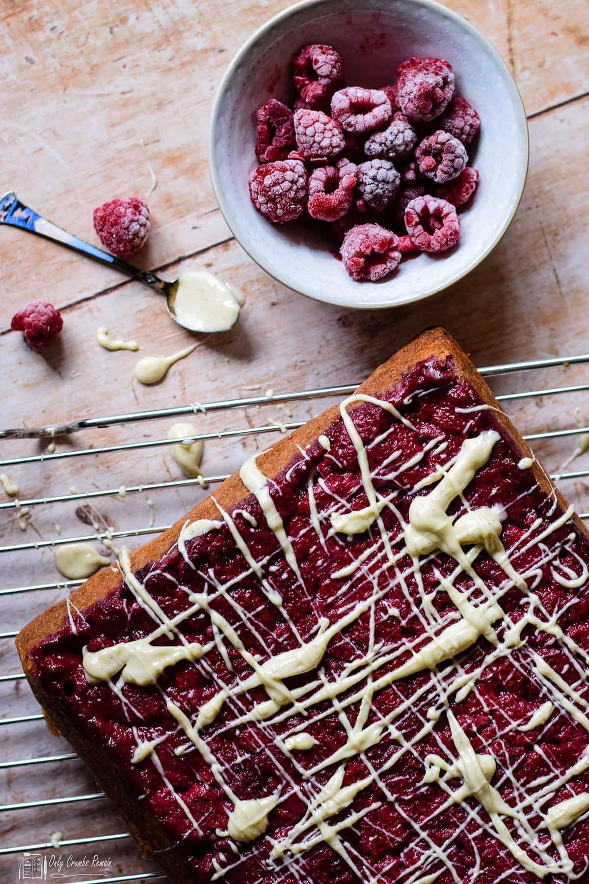 raspberry and white chocolate traybake cake on cooling rack with bowl of raspberries.