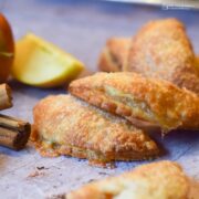 several mini apple turnovers piled on work surface with apples and cinnamon sticks next to them.