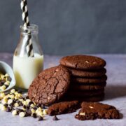 stack of easy double chocolate cookies beside a bottle of milk with 2 straws. On the other side a bow of chocolate chips spilling out in front of the milk and cookies.