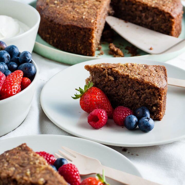 slice of sticky walnut cake with fresh berries and the rest of the cake on plate behind.