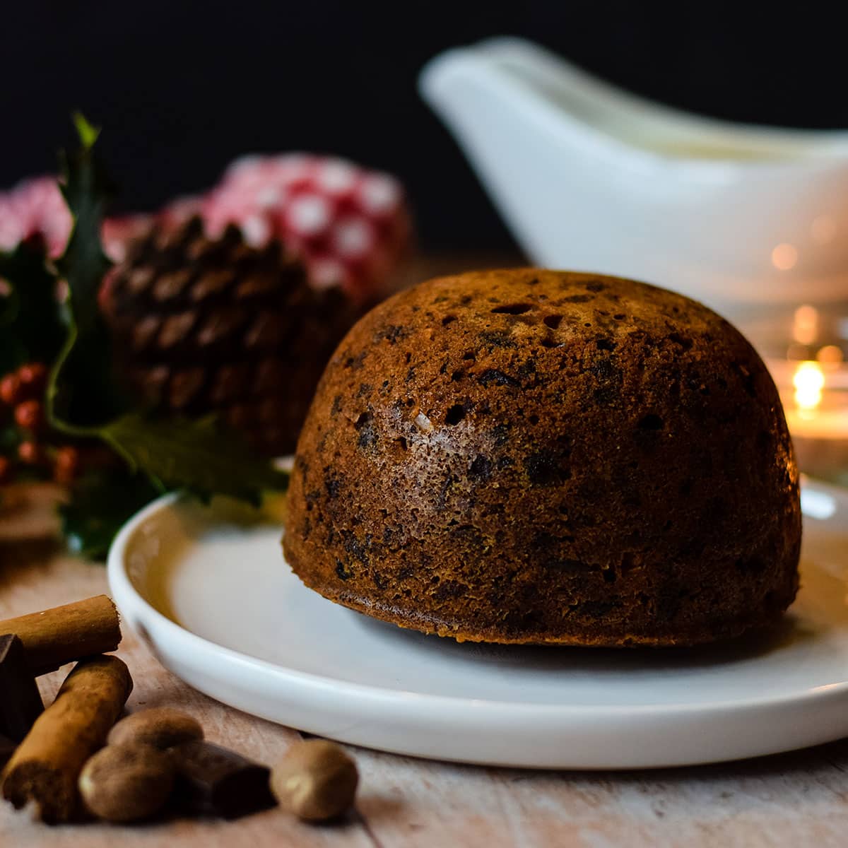 whole chocolate orange Christmas pudding on serving plate with sauce boat and Christmas decorations behind and spices to side.