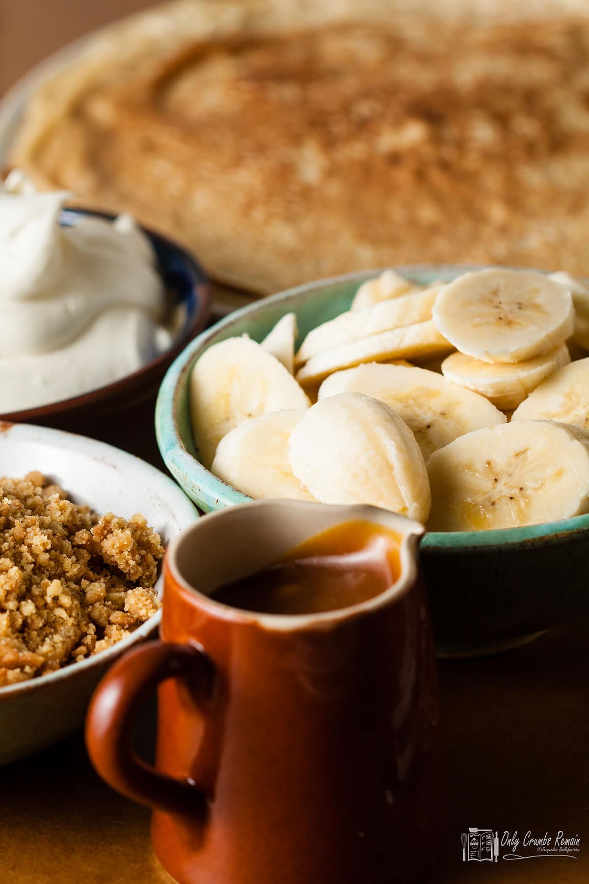 stack of pancakes in background with bowls of cream, crumbs and sliced banana withjug of caramel sauce in front.
