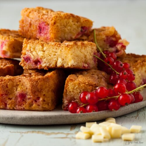 stack of redcurrant and white chocolate blondies on a plate with fresh redcurrants and white chocolate chunks on the side