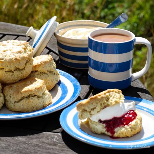 Garden table with a plate of plain scones, mug of tea and tea plate with a scone filled with jam and cream.